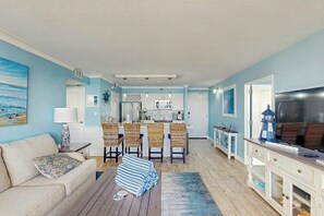 A living room with a sofa, a coffee table, and a dining area with chairs. There is a kitchen in the background with a refrigerator and a microwave. - Ocean Front Escape at the San Luis (Galveston)