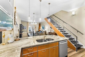 A kitchen with a marble countertop, stainless steel sink, and wooden cabinets. There is a staircase with a glass railing leading to another level. - Palm Bay - Pelican Perch (Corpus Christi)