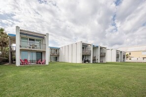 Um edifício moderno com telhado plano, varanda coberta com cadeiras vermelhas e gramado bem cuidado.