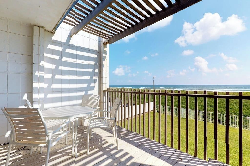 Un balcón exterior con vista al océano, una mesa y sillas blancas, y una estructura de pérgola.