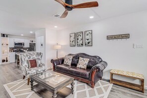 A modern living room with a leather sofa, a glass coffee table, a patterned armchair, a wooden bench, and a ceiling fan. - Corpus Christi Charm (Corpus Christi)