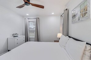A bedroom with a bed, a nightstand, a ceiling fan, a window with curtains, and a wall-mounted lamp. - Corpus Christi Charm (Corpus Christi)