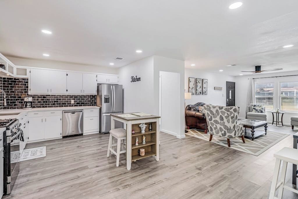 A modern kitchen with white cabinets, a built-in oven, and a refrigerator. There is a dining area with a table and chairs, and a living room with a sofa and a patterned armchair.