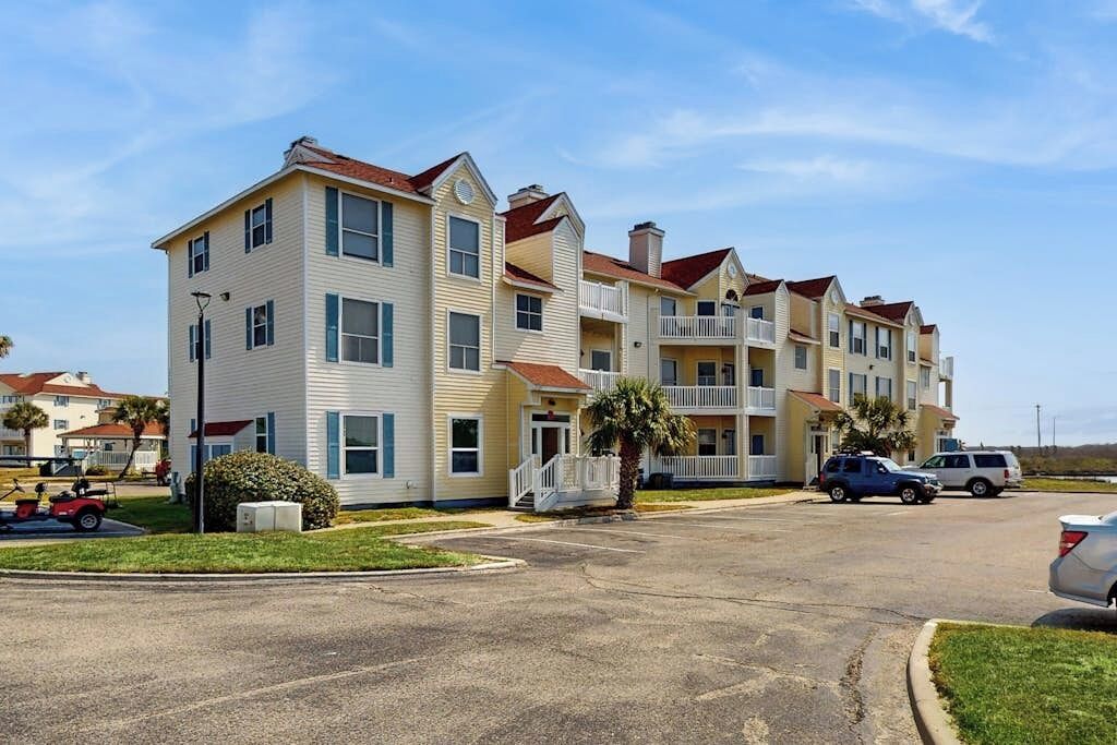 A multi-story residential building with a central entrance, balconies, and a parking area with vehicles.