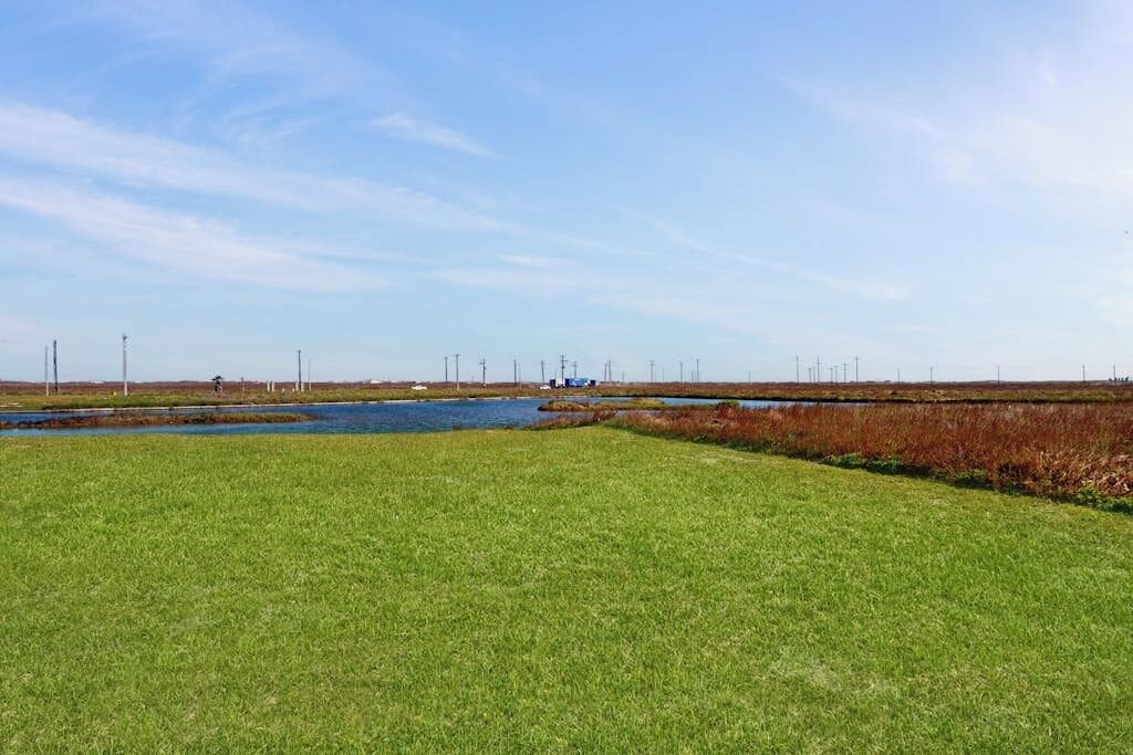 Eine flache Landschaft mit einem Gewässer, grasbewachsenen Flächen und einem Gebäude in der Ferne.