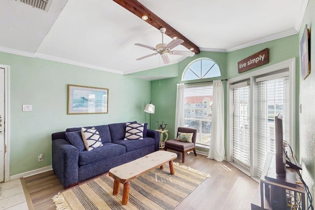 A living room with a sofa, a coffee table, a ceiling fan, a TV, and a window with blinds.