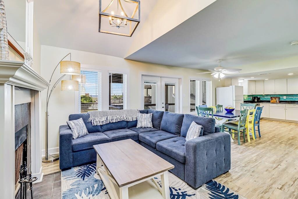 A modern living room with a sectional sofa, a coffee table, and a fireplace. There is a dining area with a table and chairs, and a kitchen in the background.