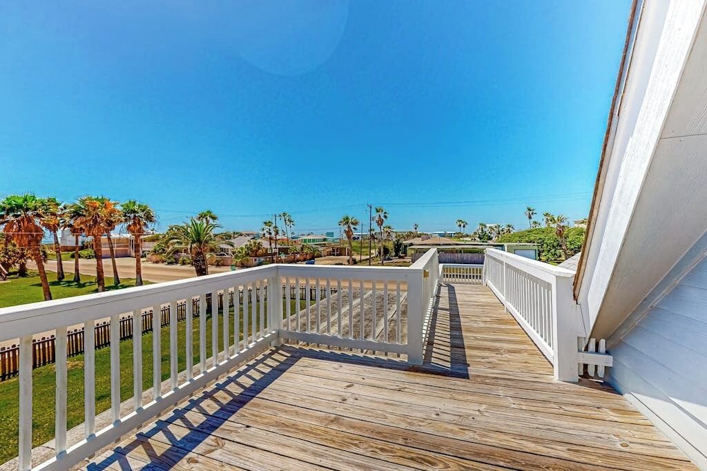 A wooden deck with white railings, leading to a view of a beach with palm trees and a clear sky.