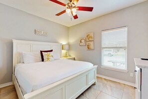 Ein Schlafzimmer mit Bett, Deckenventilator, Nachttisch mit Lampe, Fenster mit Jalousien und Wandbildern.