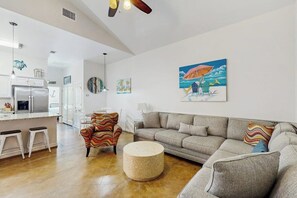 A modern living room with a grey sofa, a colorful armchair, and a round ottoman. There is a kitchen area with a refrigerator and a dining table. - Anegada Village - Dreamcatcher (Corpus Christi)