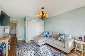 Ein Wohnzimmer mit einem Ecksofa, einem gemusterten Teppich, einer Kristallleuchtre und einem an der Wand befestigten Fernseher.