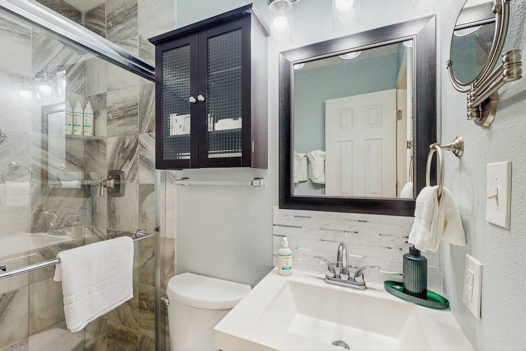 A bathroom with a glass shower enclosure, a white toilet, a sink with a green soap dish, and a mirror with a black frame.