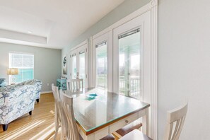 A room with a glass-top dining table, a sofa, and a view of a balcony. - Heron's Hideaway - Screened Porch, Walk to Bay (Rockport)