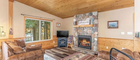 Una sala de estar acogedora con chimenea de piedra, techo de madera y grandes ventanales con vistas a un bosque.