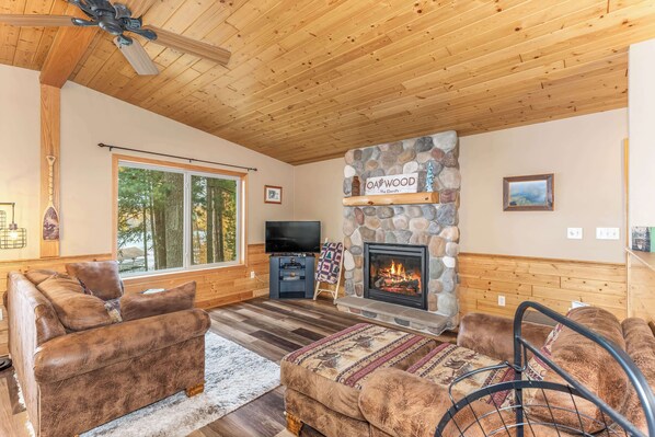 A cozy living room with a stone fireplace, wooden ceiling, and large windows overlooking a forest.