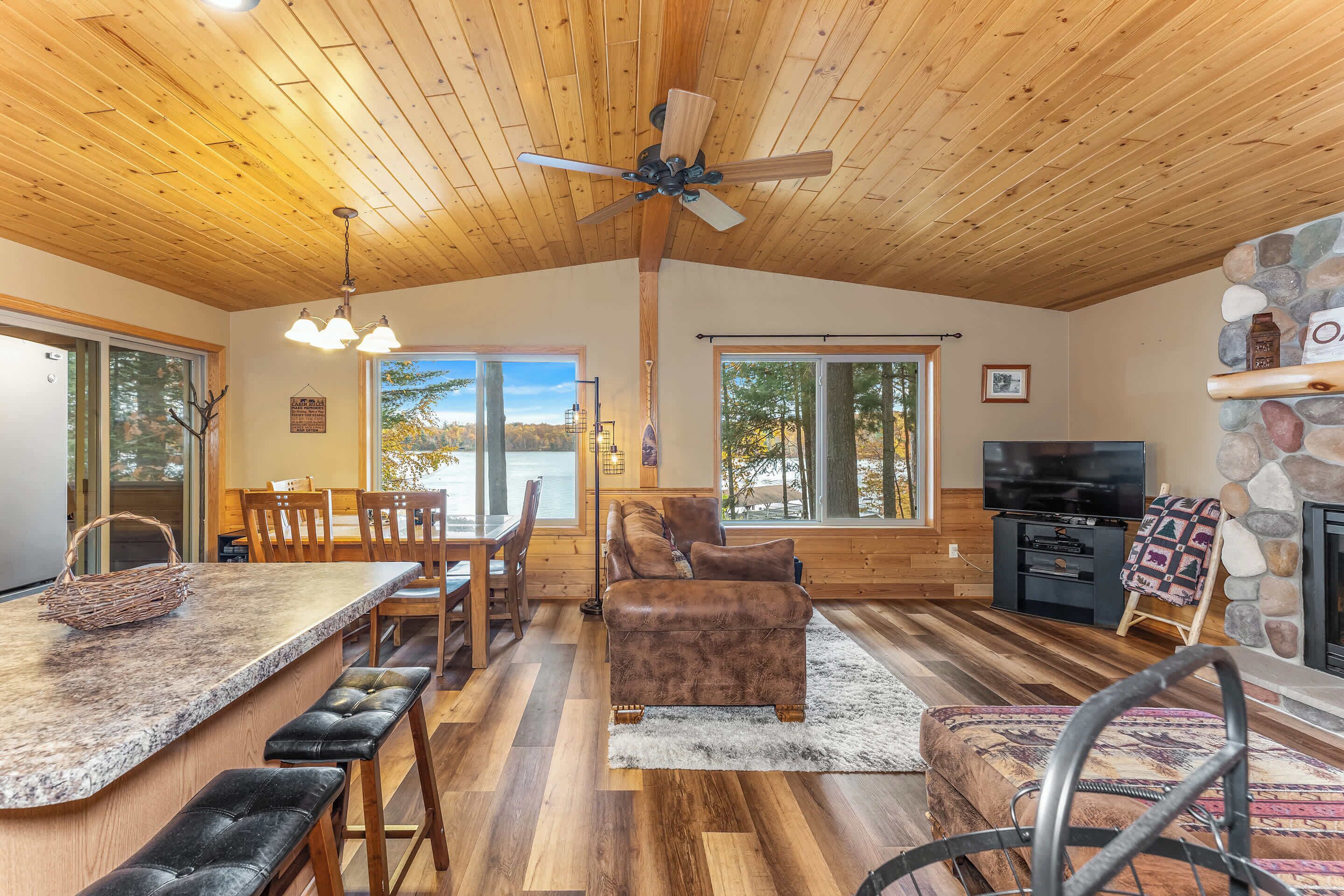 A spacious living area with a wooden ceiling, a stone fireplace, a television, and a large window offering a view of trees.