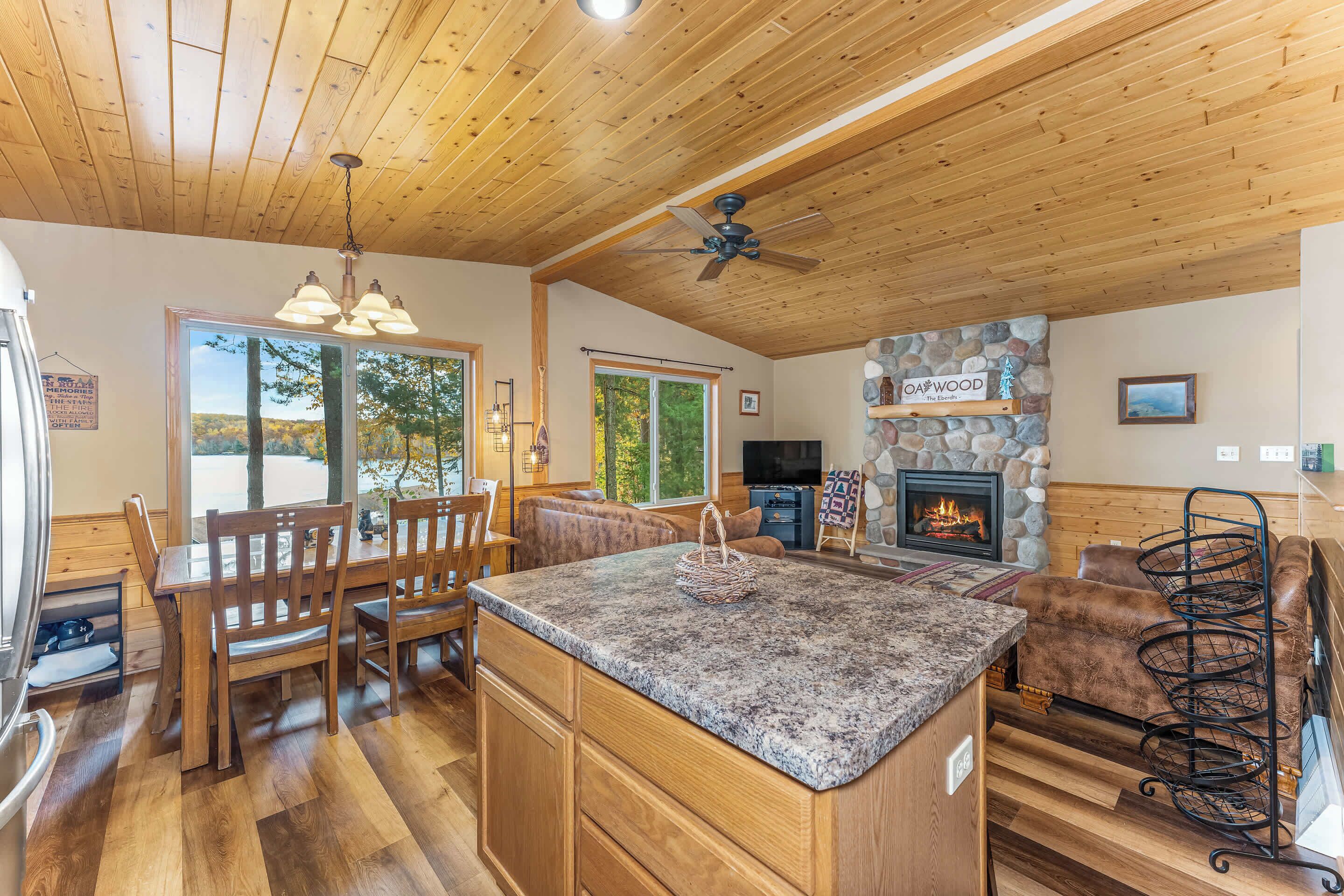 Amplia sala de estar con chimenea de piedra, techo de madera y un ventanal con vistas a un bosque.