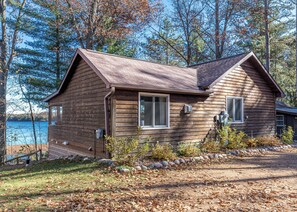 A wooden cabin with a sloped roof, a window, and a door, situated by a lake with trees in the background. - Pinewood Cabin, Resort, Lake, Beach, Kayaks & More (Lac du Flambeau)