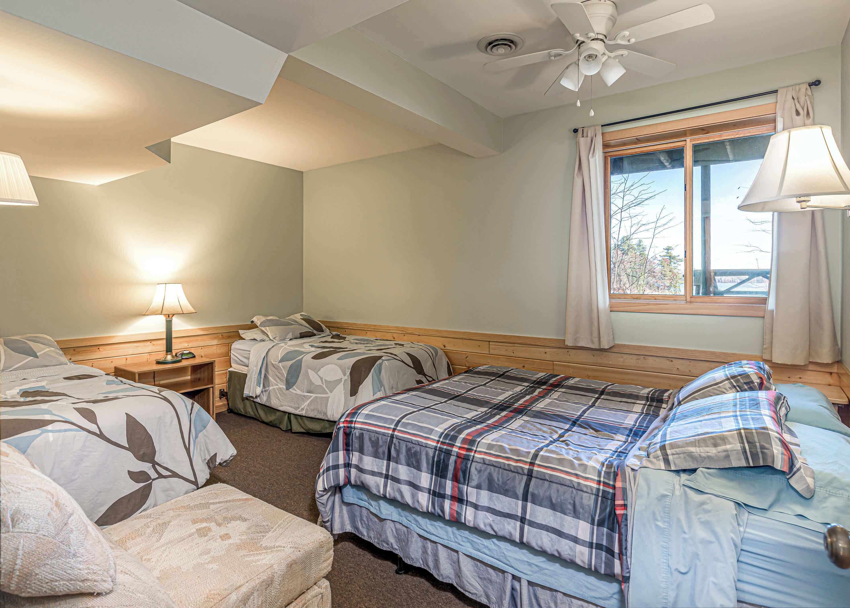 A bedroom with two beds, a ceiling fan, a window with curtains, and a bedside table with a lamp.