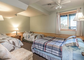 Quarto com duas camas, ventilador de teto, janela com cortinas e mesa de cabeceira com abajur.