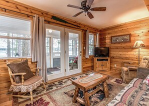 A cozy cabin interior with a wooden interior, a ceiling fan, a television, and a fireplace.