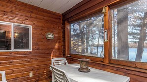 Interior de una cabaña con pared de madera, una mesa con sillas, un ventanal con vistas a árboles y un lago, y una campana en la pared.
