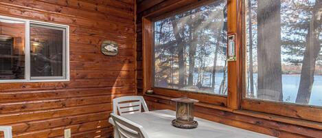 Interior de una cabaña con pared de madera, una mesa con sillas, un ventanal con vistas a árboles y un lago, y una campana en la pared.