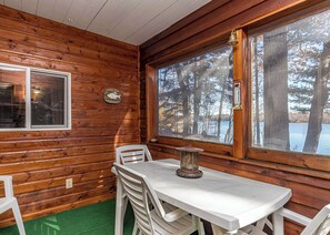 Interior de cabana com parede de madeira, mesa com cadeiras, janela com vista para árvores e um lago, e um sino na parede.