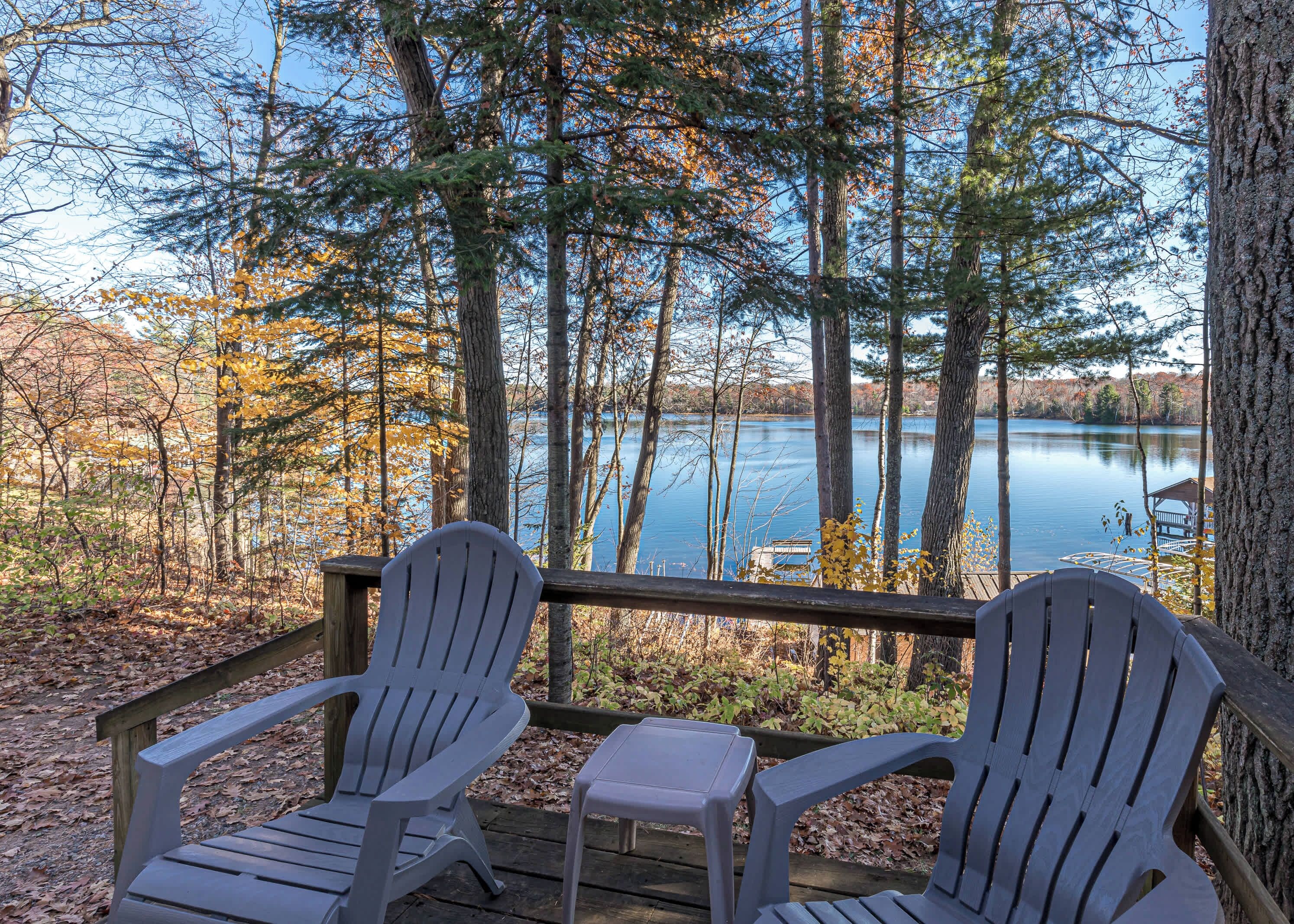 Una terraza con dos sillas Adirondack y una mesita, con vista a un lago tranquilo rodeado de árboles.