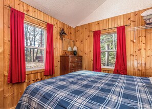 A bedroom with wooden walls, a plaid bedspread, and red curtains.