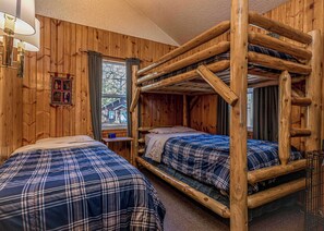 Quarto estilo cabana com duas beliches, paredes de madeira, uma janela com cortinas e uma pequena mesa.