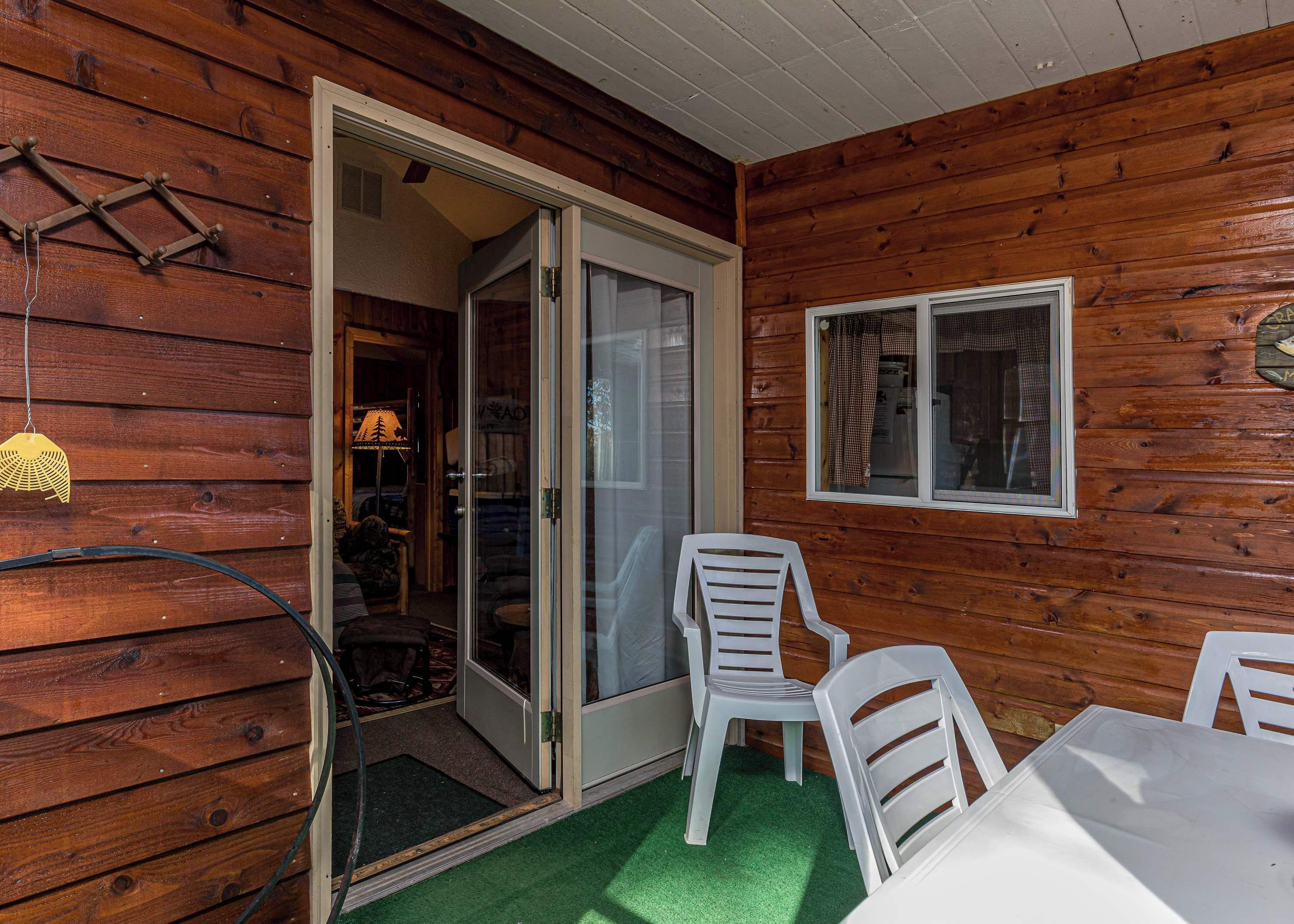 A wooden cabin with a sliding glass door leading to an interior space, a white plastic chair, and a table with a green mat.