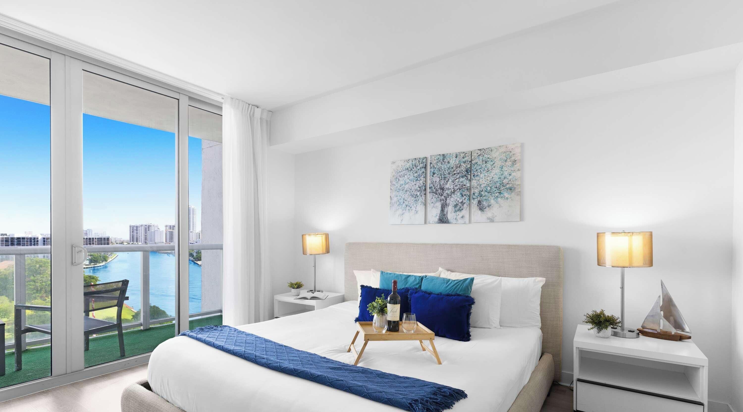 A hotel room with a large bed, two bedside tables, a view of a cityscape and water through floor-to-ceiling windows, and a painting on the wall.