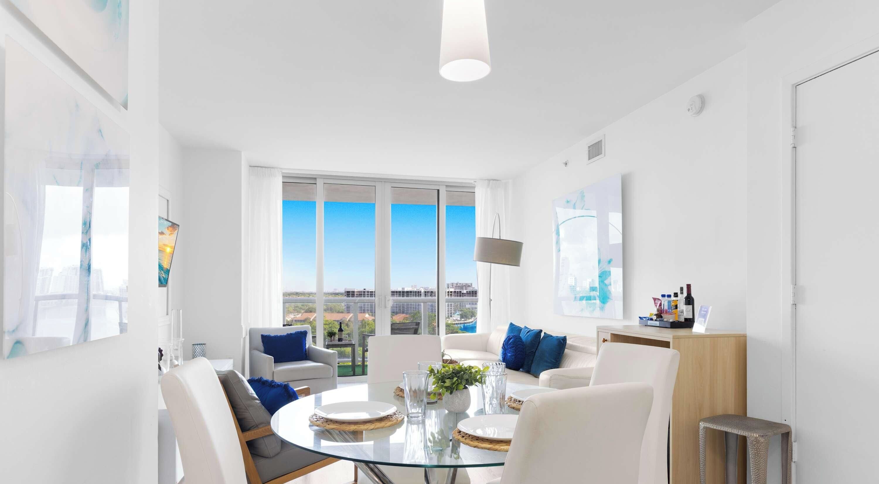 A modern living room with a glass dining table, white sofas, and a large window offering a city view.
