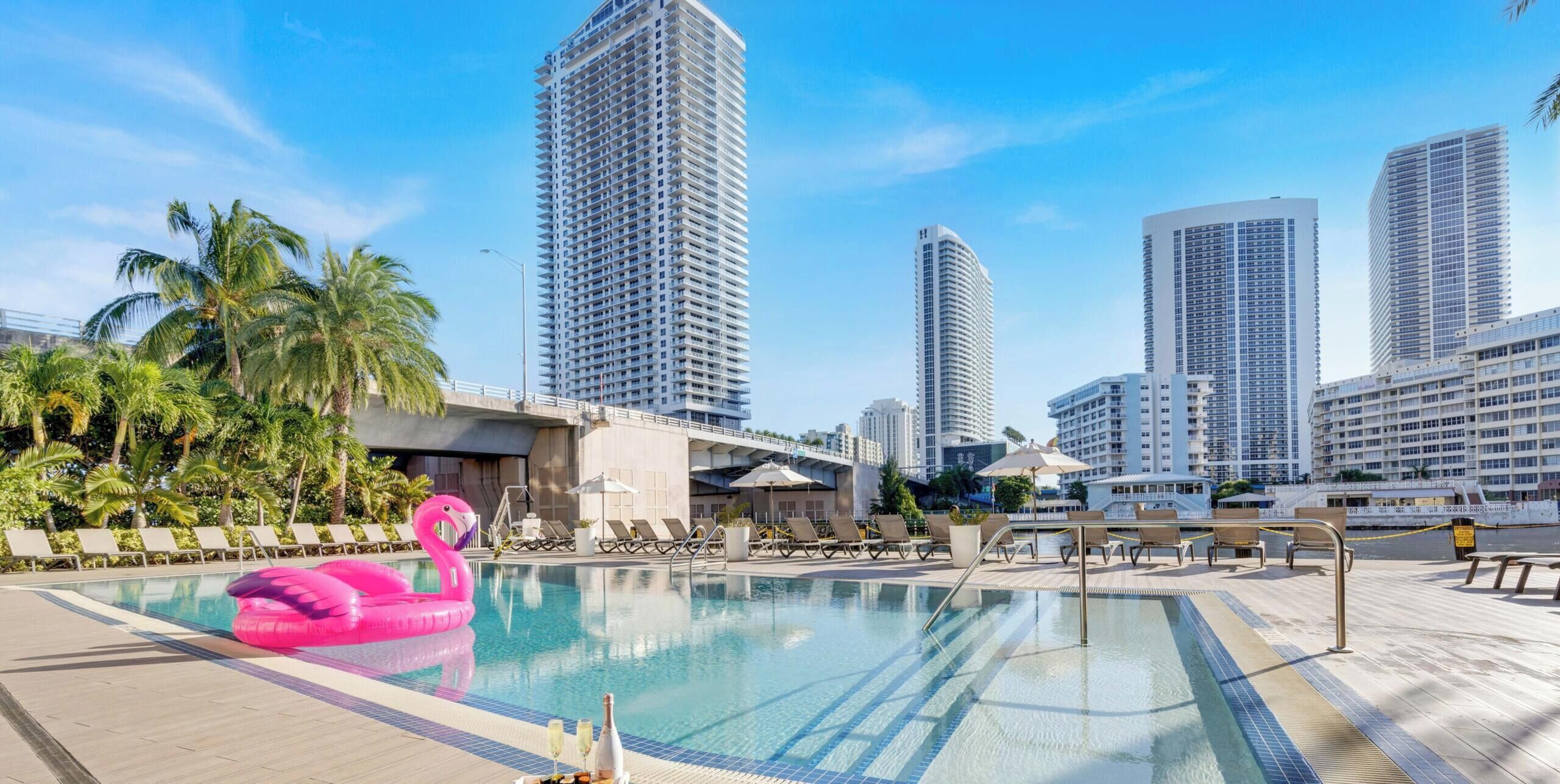 A swimming pool with lounge chairs and a pink flamingo float, surrounded by tall buildings and palm trees.