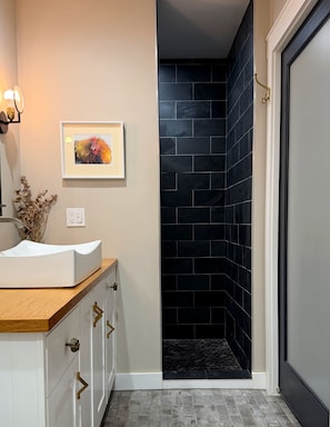 Un baño con tocador blanco, un cuadro enmarcado y zona de ducha con azulejos negros.