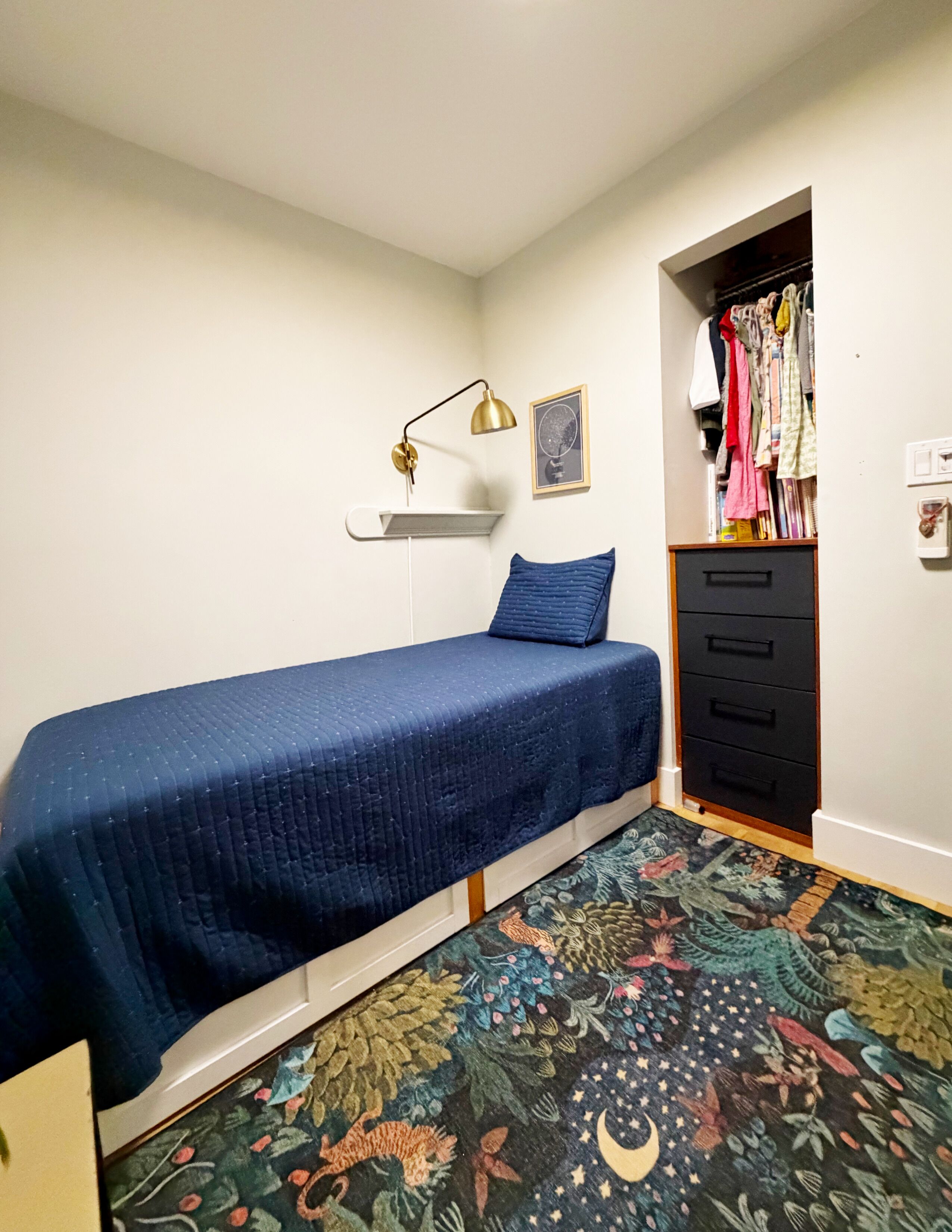 Une petite chambre à coucher, bien rangée, avec un lit simple, une lampe de chevet, une commode et un placard.