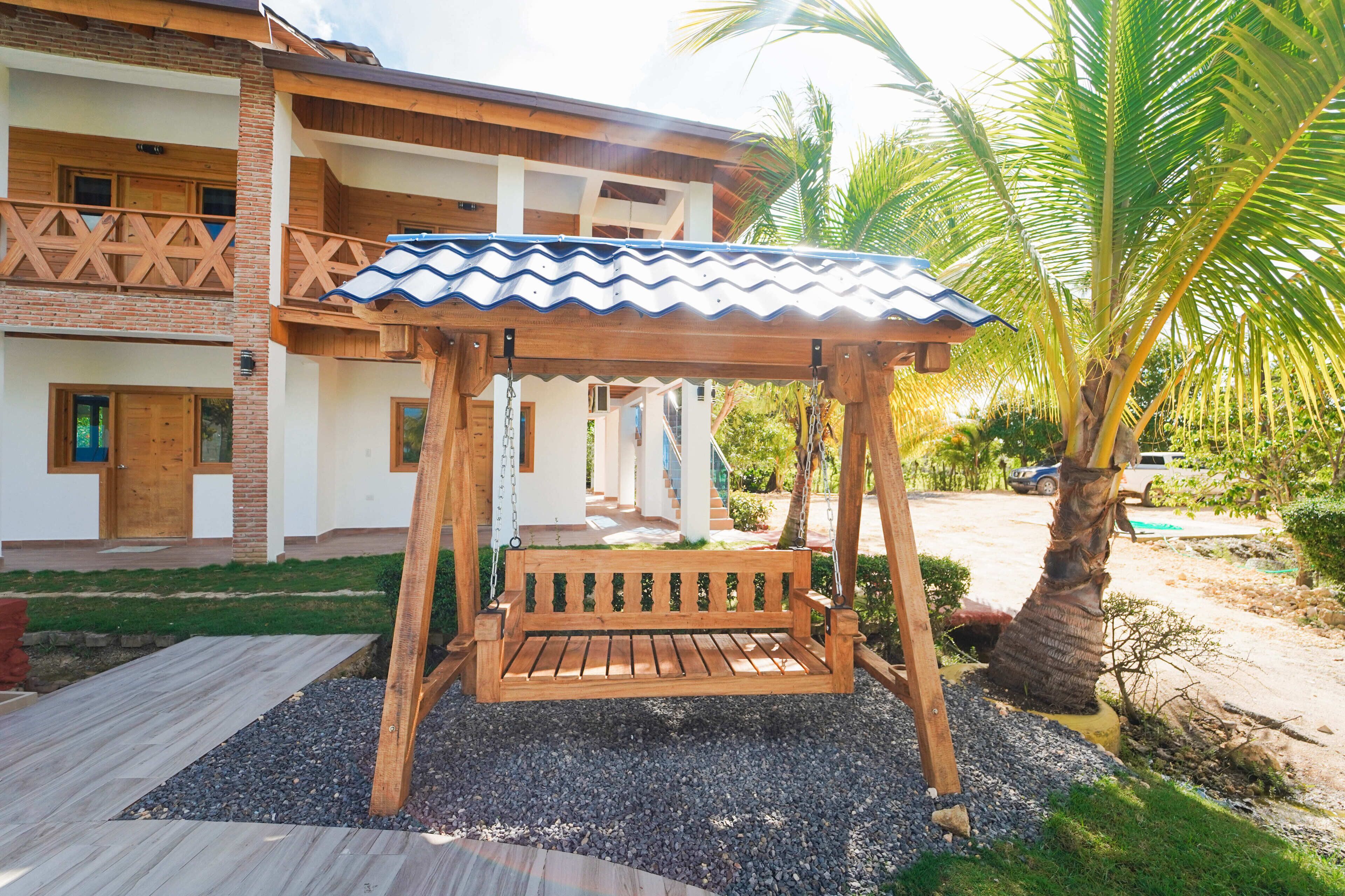 A wooden swing set with a metal roof, situated in a garden area with a paved walkway and a building in the background.