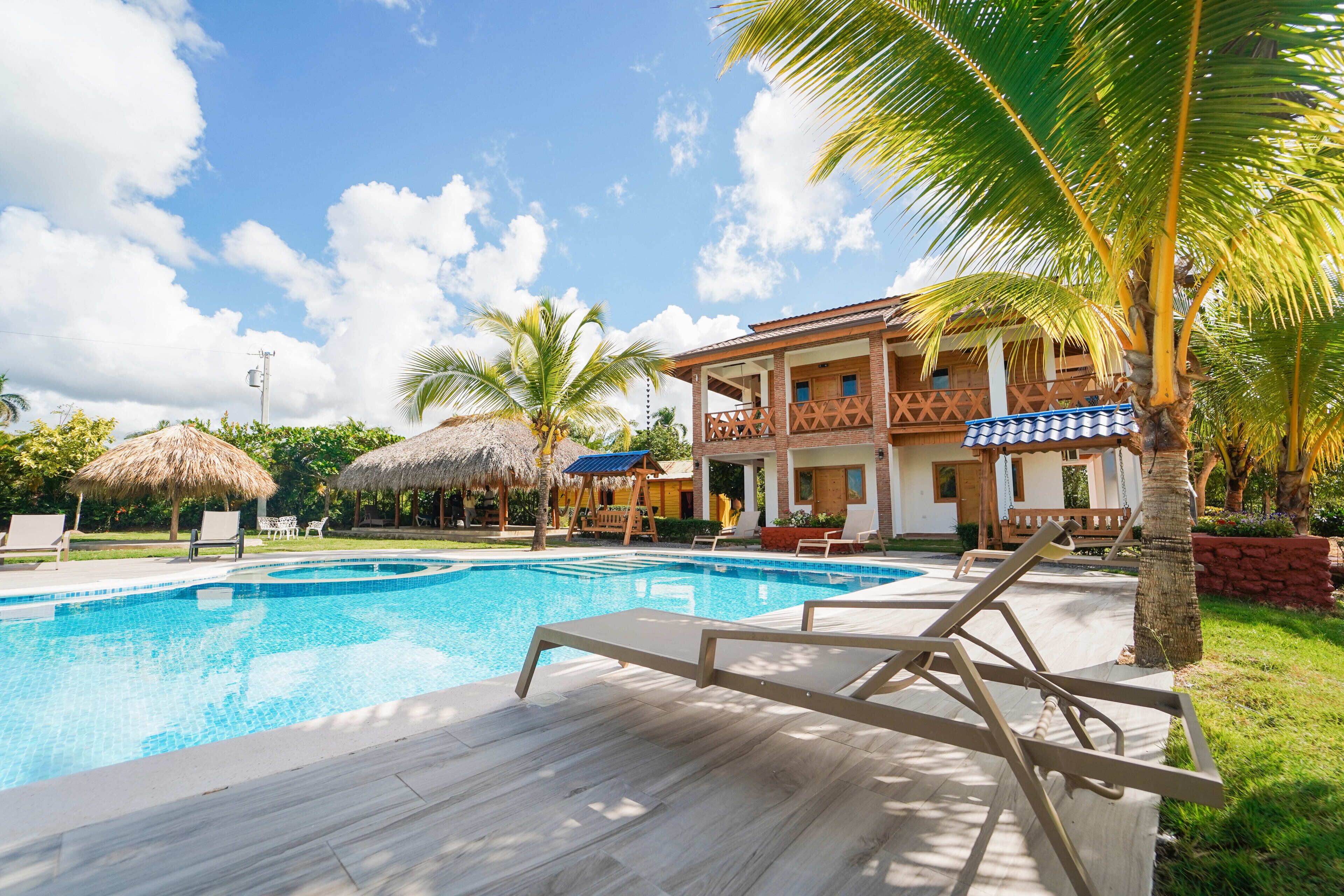 A swimming pool with lounge chairs, a thatched-roof structure, and a multi-story building with balconies.