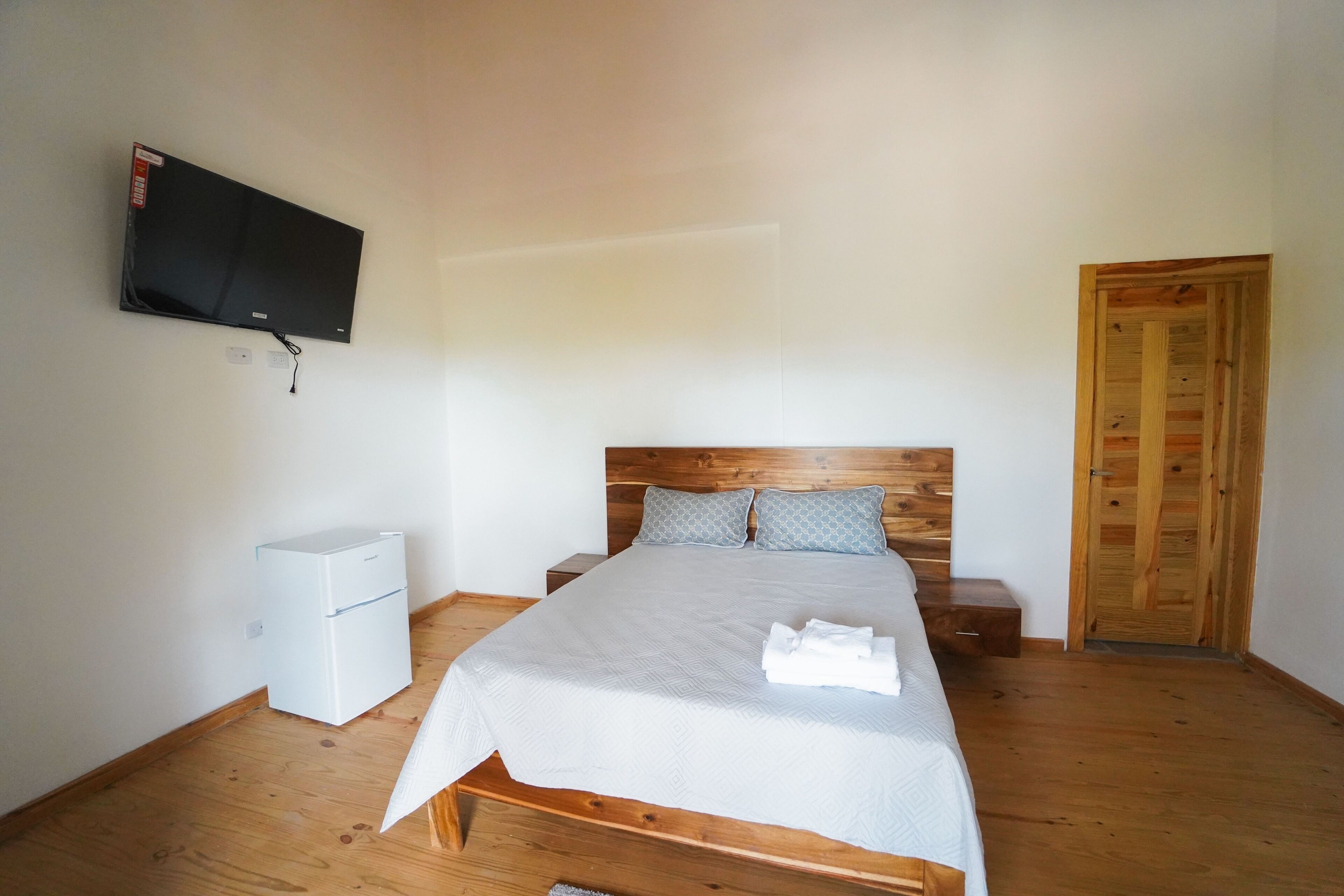 Un dormitorio con una cama, un televisor montado en la pared, un armario de madera y una mesita de noche blanca.