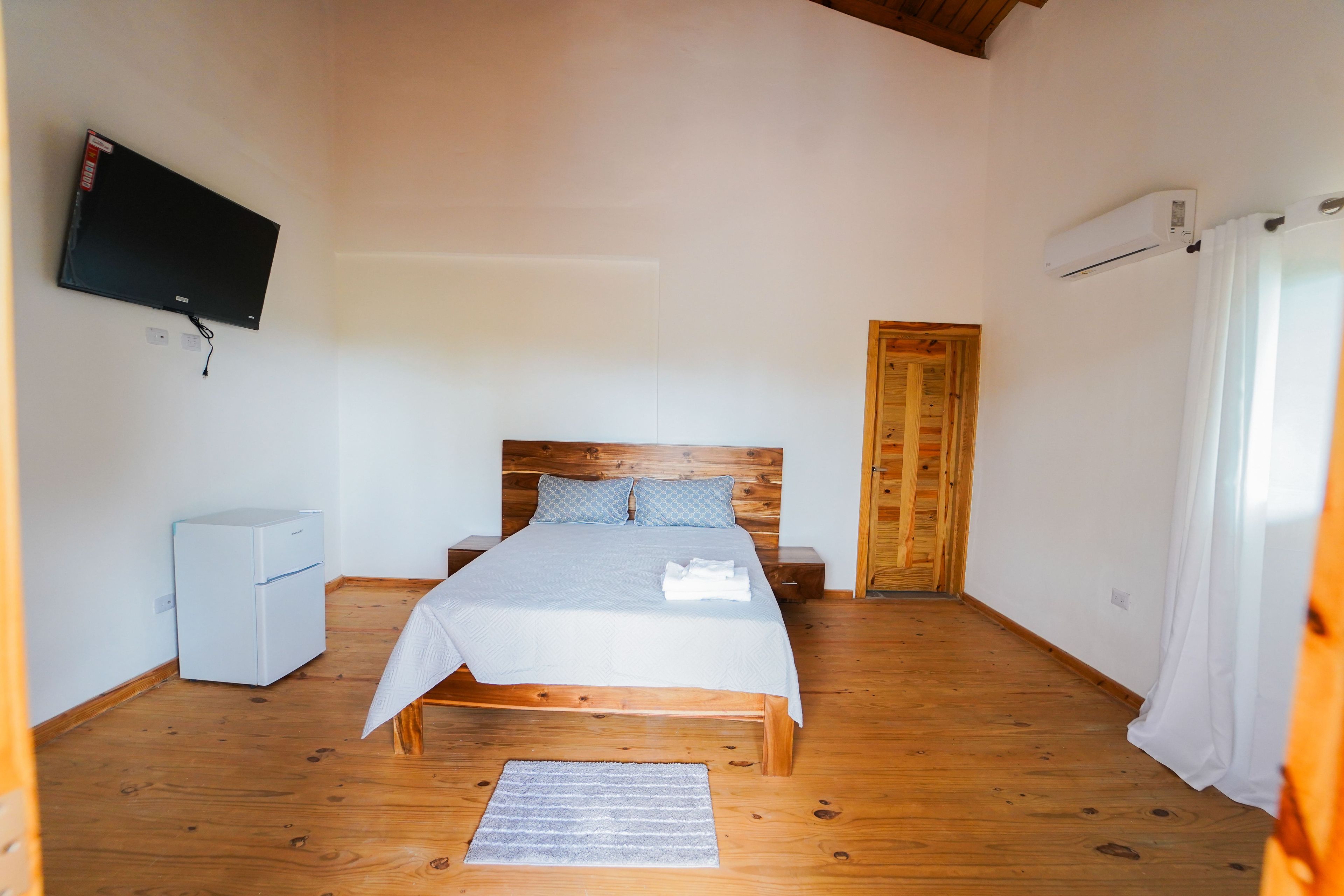 A bedroom with a wooden bed, a small white refrigerator, a television on the wall, and a wooden door.