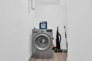 A compact, modern laundry room with a front-loading washing machine, a blue detergent bottle, and a vacuum cleaner. - 3BHK Close to Downtown/Whyte Ave/UOFA (Edmonton)