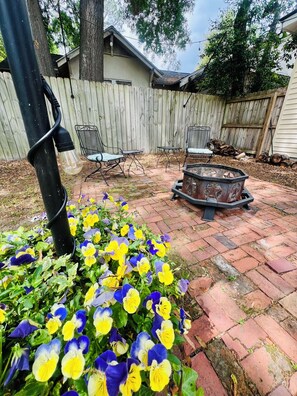 Un giardino sul retro con un patio in mattoni, una recinzione in legno, un braciere in metallo e diversi tipi di fiori.