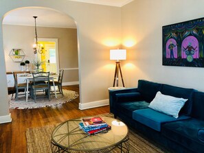 A living room with a blue sofa, a glass coffee table with books, a lamp, and a painting on the wall. - "Music & Martinis" Charming Cottage Near Medical District (Augusta)