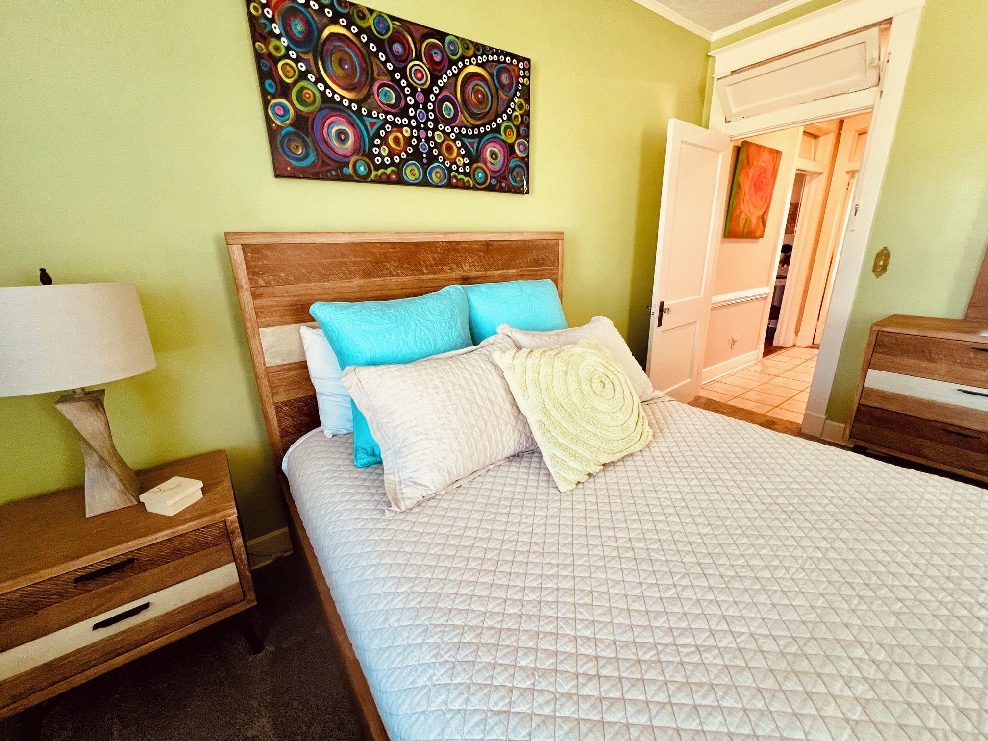 Habitación de hotel con cama, mesita de noche, lámpara, un cuadro en la pared y una puerta que da a otra habitación.