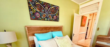 A hotel room with a bed, a nightstand, a lamp, a painting on the wall, and a door leading to another room.