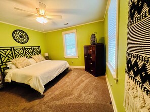 Una camera da letto con un letto, una cassettiera, una decorazione a parete, una finestra e una ventola a soffitto.