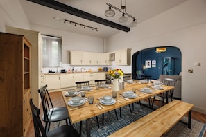 Un comedor con una mesa de madera dispuesta para cuatro personas, rodeada de sillas, en una habitación con una cocina al fondo y una pared azul decorativa.