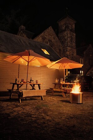 An outdoor dining area with wooden tables and benches, covered by large umbrellas, and a fire pit in the center. - Historic Clocktower House | Spa Barn | Sleeps 12 (Cumberland)