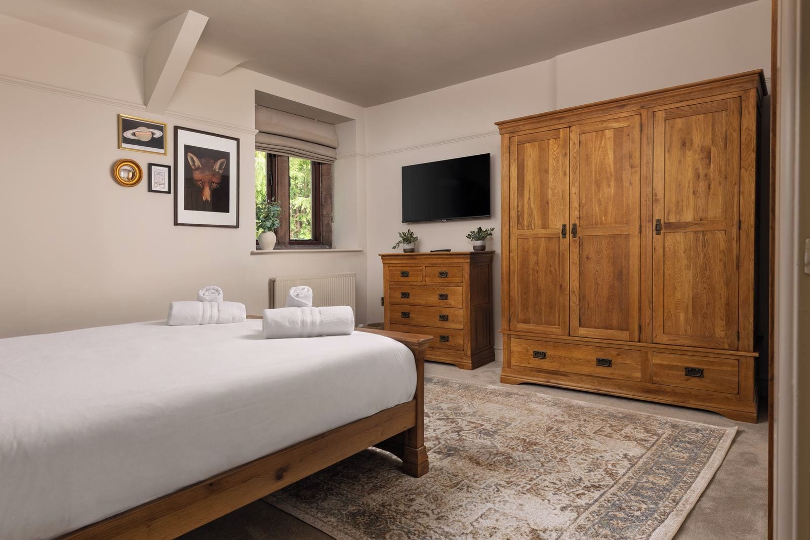 A bedroom with a large bed, wooden wardrobe, a television, and a window with a view of greenery.
